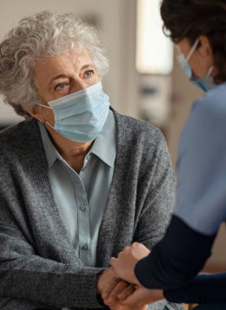 Elderly woman talking with a doctor while holding hands at home and wearing face protective mask. Worried senior woman talking to her general pratictioner visiting her at home during virus epidemic. Doctor explaining about precautionary measures during virus pandemic to old lady and takes care of her.